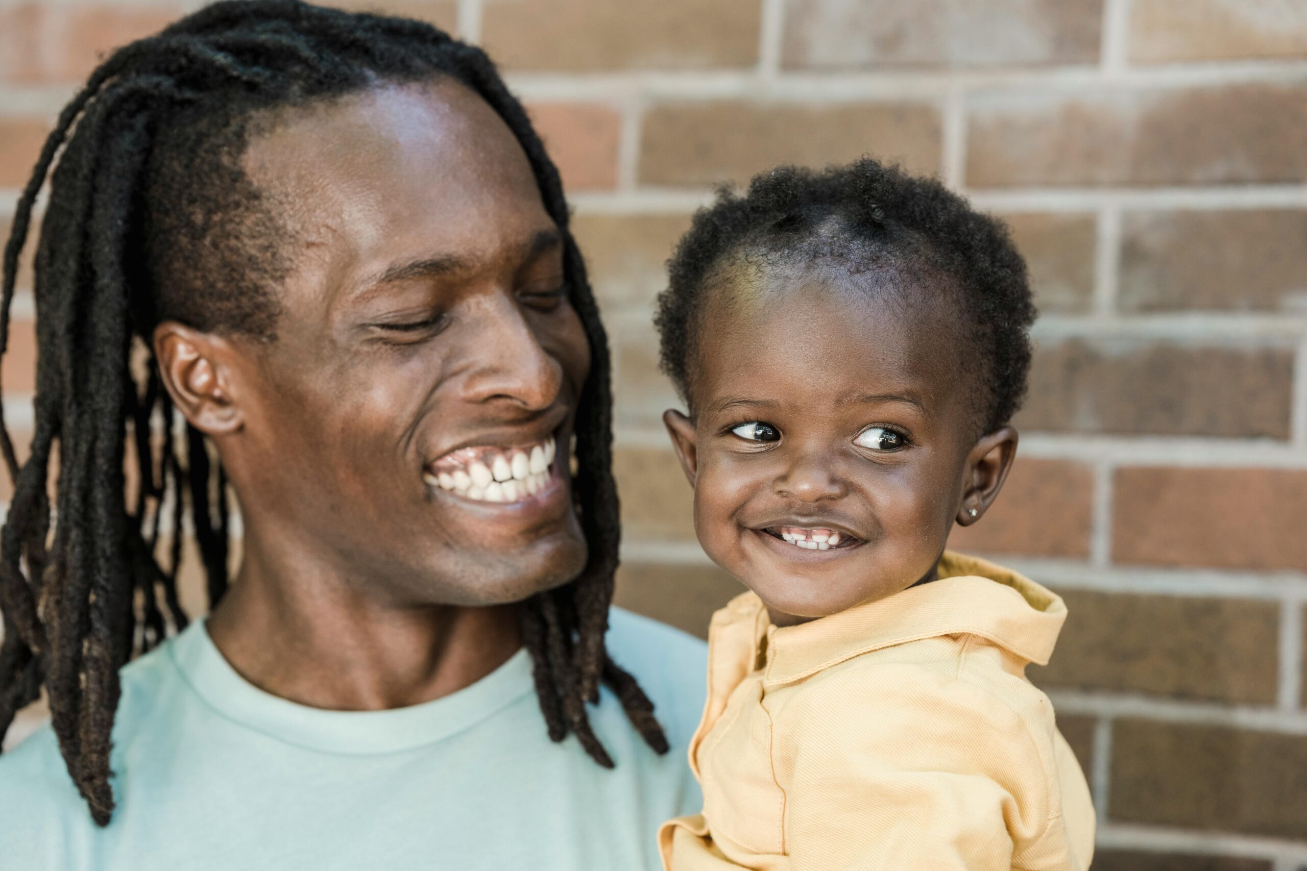 A father with dreadlocks joyfully holding his smiling young daughter against a brick wall backdrop.